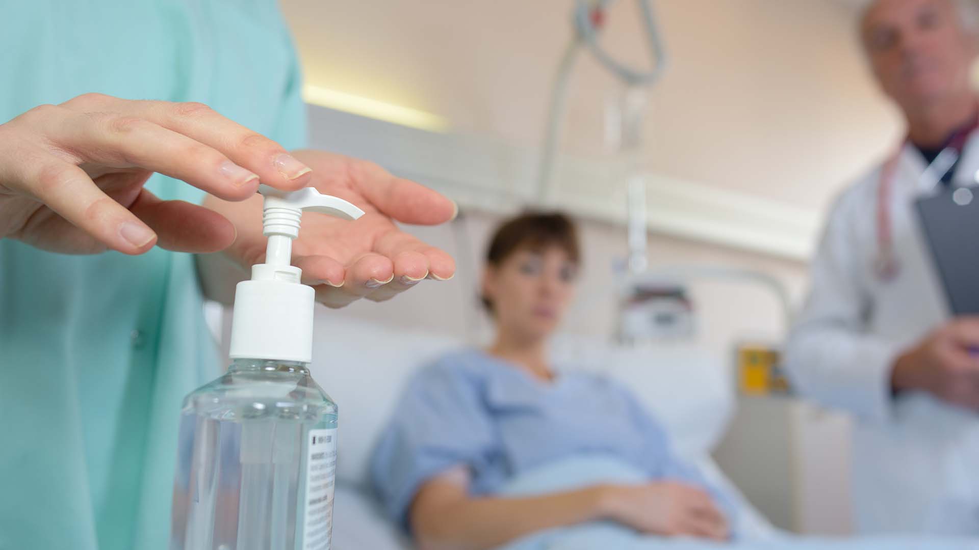 Eine Krankenschwester desinfiziert sich die Hände. Im Hintergrund ist eine Patientin in einem Bett liegend zu sehen.